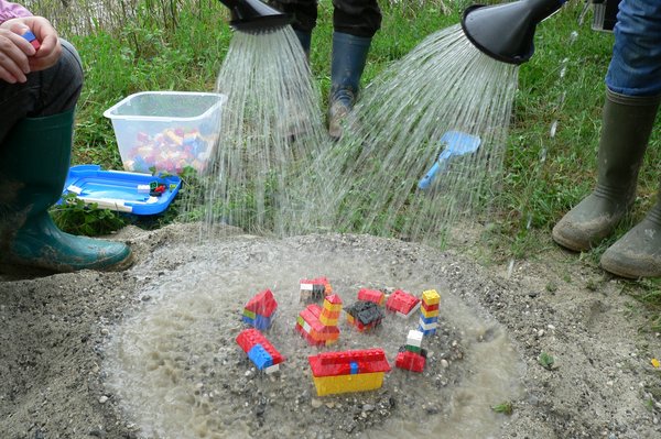 mehrere Personen gießen auf Spielfiguren in einem Sanskasten