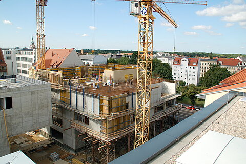 Auf dem Bild ist ein Wohnhaus im frühen Baustadium zu sehen. Im Bildvordergrund und -hintergrund befinden sich Kräne.