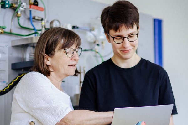 Zwei Personen stehen vor einer technischen Wandinstallation. Eine zeigt auf den Laptop der anderen, beide tragen Brillen und wirken konzentriert.