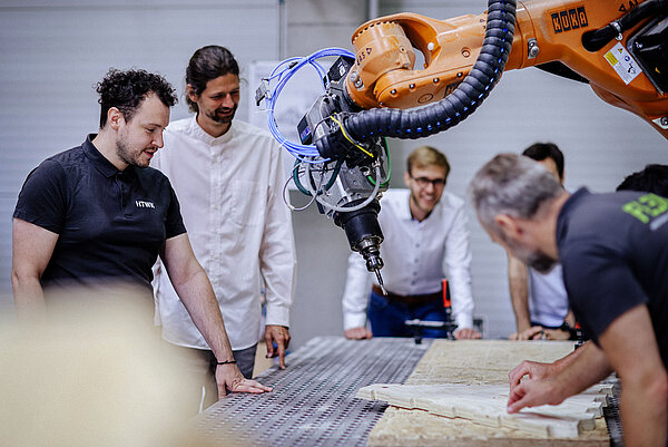 TimberBot mehrere Männer stehen am Werktisch um einen Industrieroboter herum. Auf dem Tisch liegen Holzbretter.