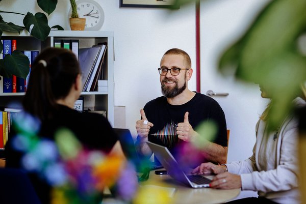 Ein Mann mit Brille sitzt an einem Tisch und zeigt Daumen hoch, während er mit zwei Personen spricht. Im Hintergrund sind Bücherregale und Pflanzen zu sehen. 