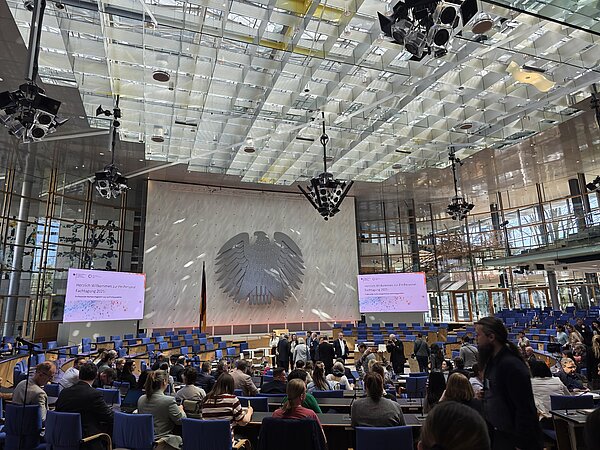 Auftakt der FH-Personal Fachtagung 2025 im ehemaligen Bundestag Großer Konferenzsaal mit Bundesadler an der Wand, zwei Leinwänden mit Begrüßungstext und vielen sitzenden sowie stehenden Menschen.