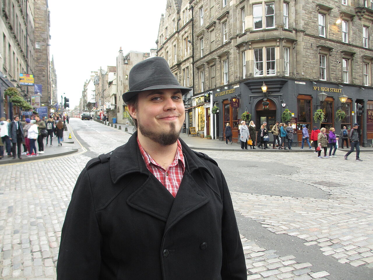 Aron Schaub mit Hut unterwegs an einer typisch britischen Straßenkreuzung vor einem Eingang zum Pub.