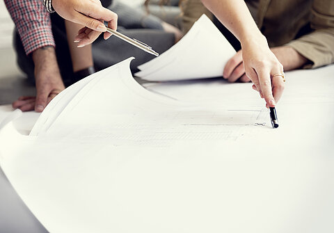 Es ist ein großes Blatt Papier zu sehen. Die Hände von zwei Personen, welche mit einem Stift auf die Skizze zeigen, sind zu sehen.