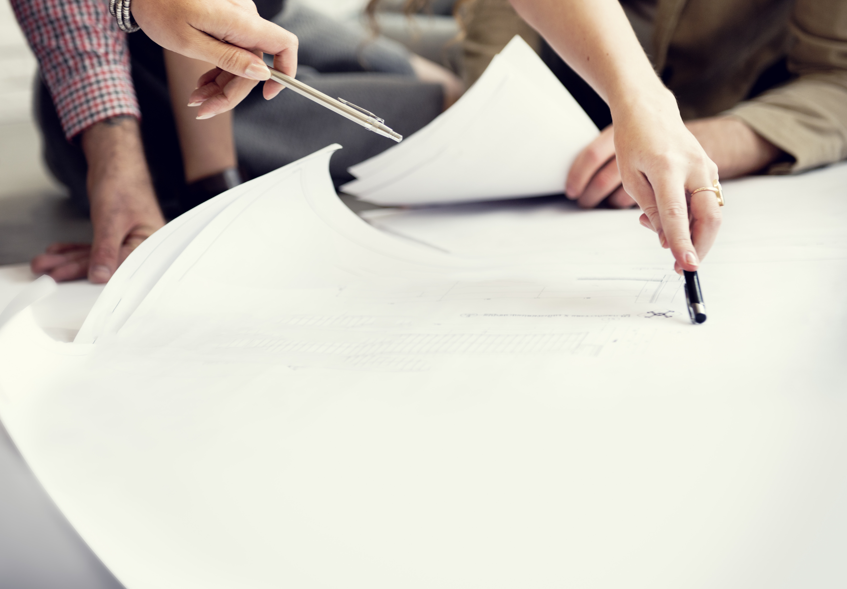 Bauwesen Planung Es ist ein großes Blatt Papier zu sehen. Die Hände von zwei Personen, welche mit einem Stift auf die Skizze zeigen, sind zu sehen.